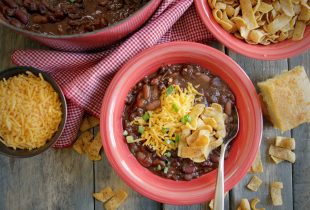 a bowl of 3 bean vegetarian chili next to frito chips, shredded cheese and a dutch oven of beans