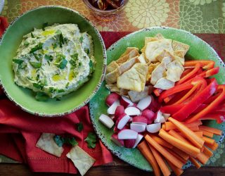 a bowl camellia bran beans hummas next to a plate of pita bread, carrots, and orange bell peppers