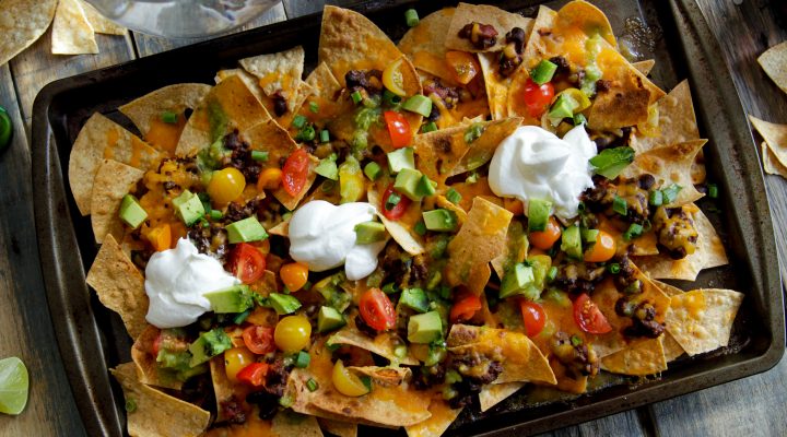 a baking pan of nachos topped with beans, sour cream, avacados and tomatos