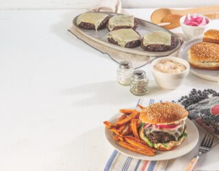 table with white tablecloth has plate of cooked Black Bean burger patties with melted cheese on them behind some condiments and salt and pepper shakers. In front of all is a circular, white, plate with cooked sweet potato fries and a completely assembled black bean burger on a toasted bun