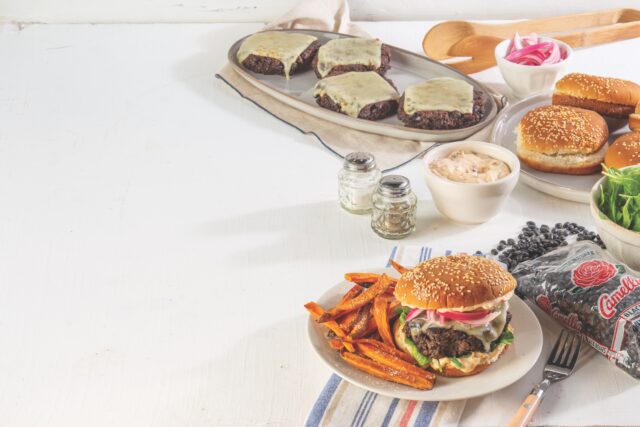 table with white tablecloth has plate of cooked Black Bean burger patties with melted cheese on them behind some condiments and salt and pepper shakers. In front of all is a circular, white, plate with cooked sweet potato fries and a completely assembled black bean burger on a toasted bun