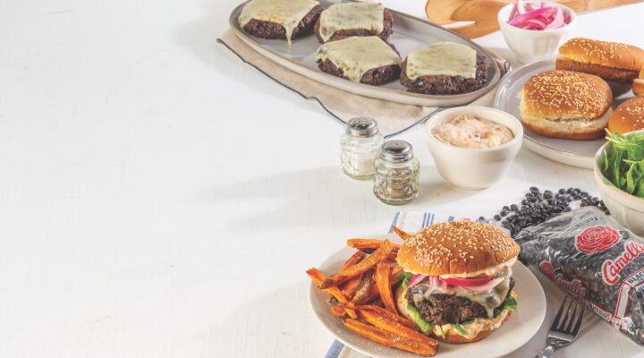 table with white tablecloth has plate of cooked Black Bean burger patties with melted cheese on them behind some condiments and salt and pepper shakers. In front of all is a circular, white, plate with cooked sweet potato fries and a completely assembled black bean burger on a toasted bun