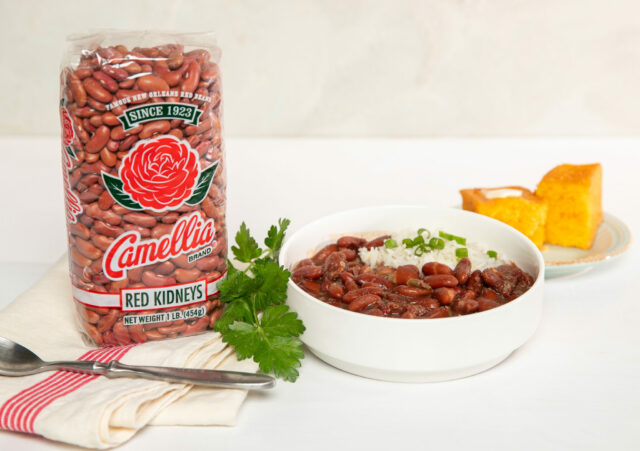 A bowl of red beans and rice garnished with green onions sits next to a bag of Camellia brand red kidney beans and cornbread on a small plate. A sprig of parsley and fork are placed beside the bowl.