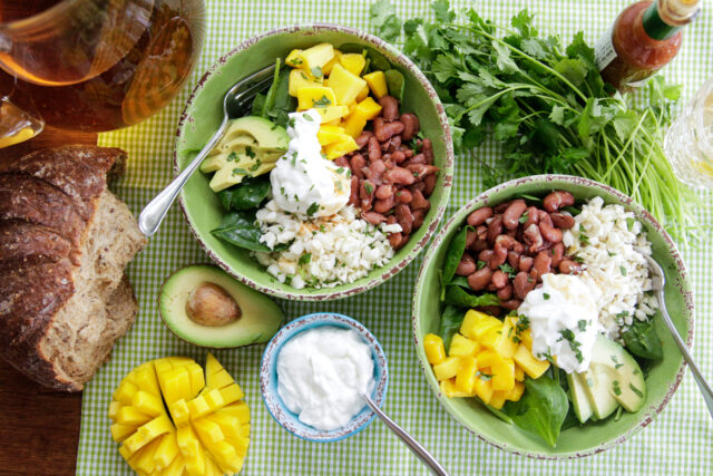 Two bowls of salad containing mango, avocado, beans, rice, and greens, topped with sour cream. Accompanied by a bowl of sour cream, sliced mango, avocado half, bread, and fresh herbs on a checkered cloth.