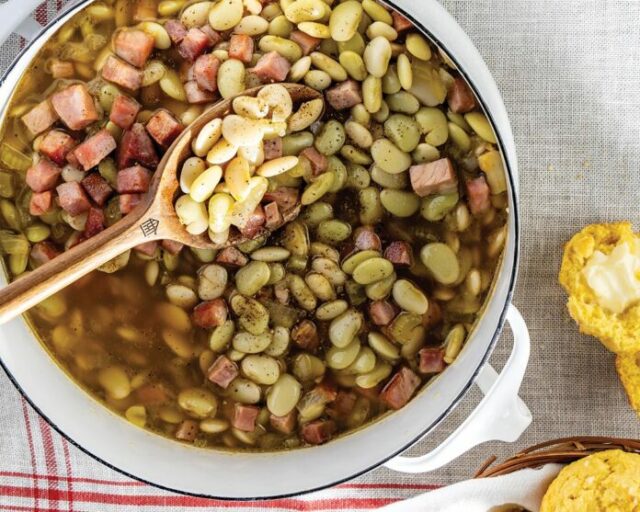 overhead image of large, white, stovetop pot on white table with white and red cloth napkin underneath and cooked butter beans inside pot