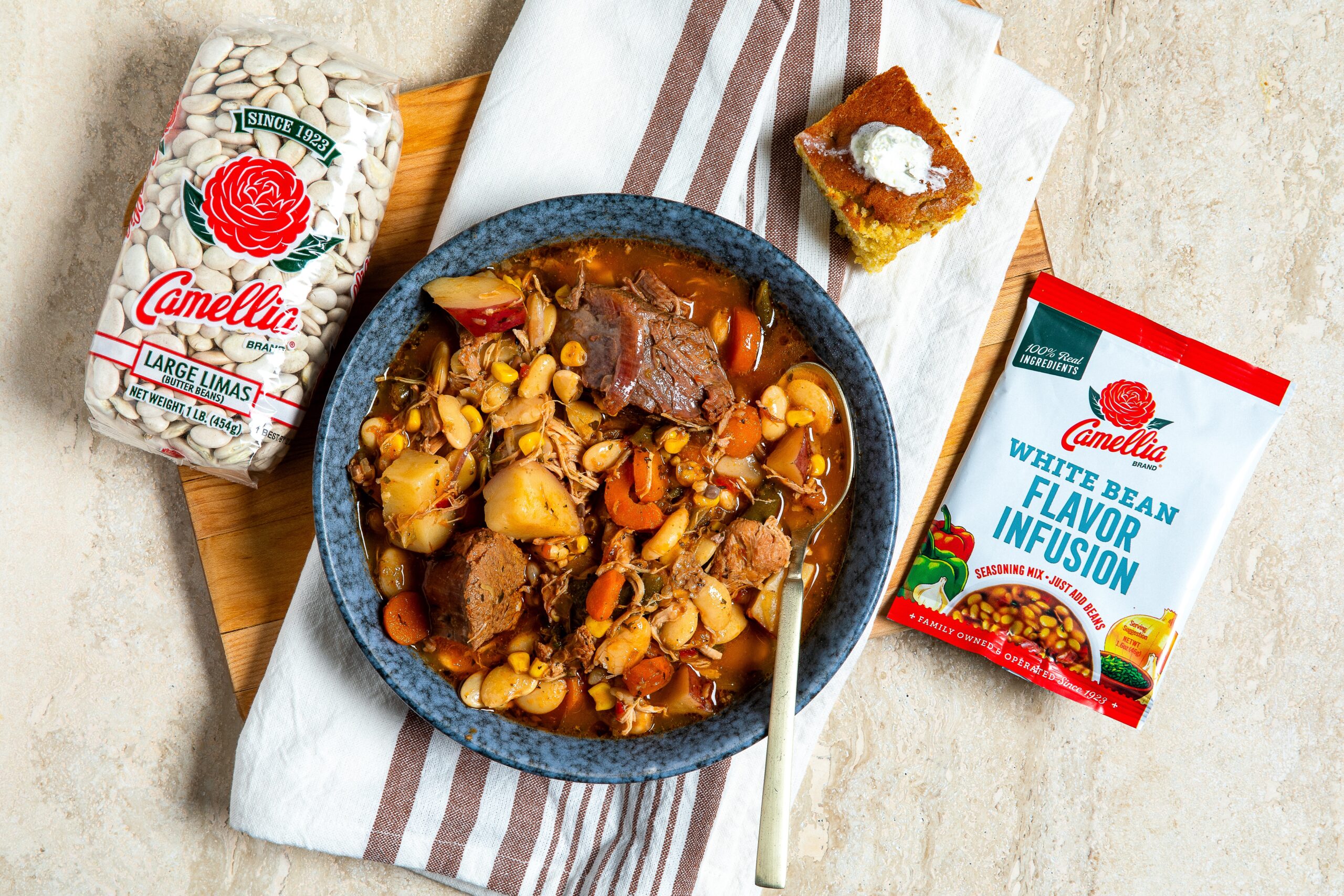 overhead image of bowl of cooked Kentucky Burgoo stew, with bag of Large Lima beans and White Bean Flavor Infusion pouch laying next to it on a light table, white napkin with centered, narrow stripes underneath