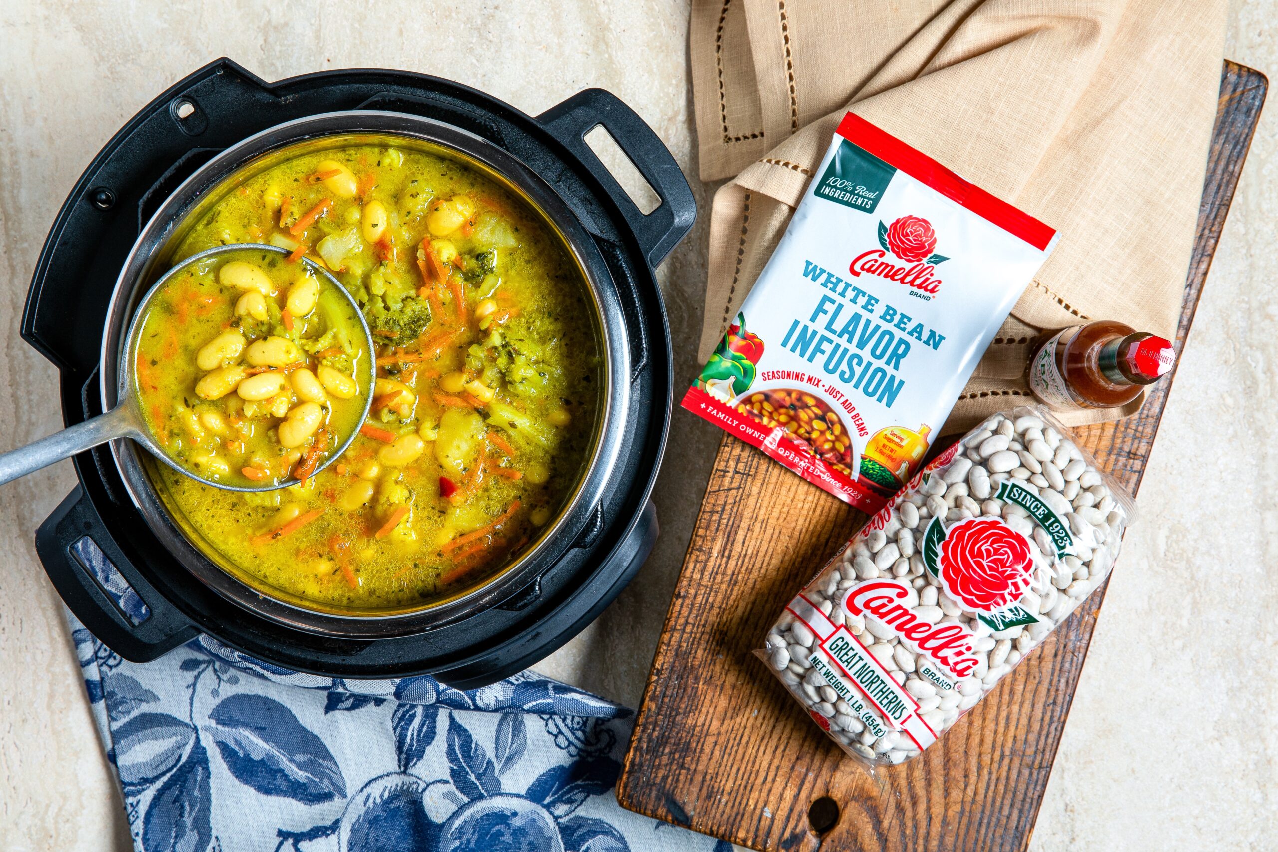 overhead image of Instant Pot full of cooked Instant Pot White Bean, Cauliflower & Broccoli Soup, with bag of Great Northern beans and Flavor Infusion pouch laying next to it on a wooden cutting board. 2 linen napkins underneath all.