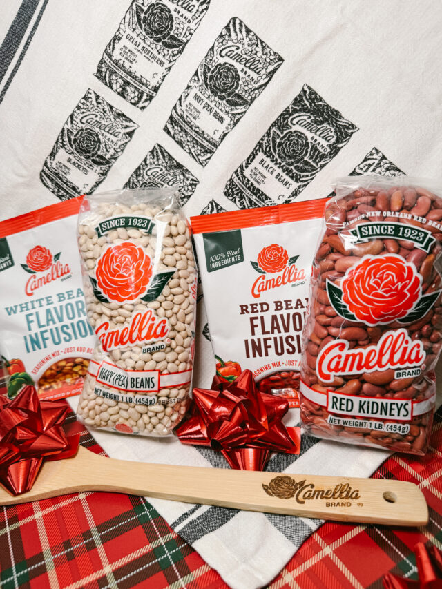 red, green, and white plaid tablecloth, with 1 pound bags of red kidneys and white navy beans, a bag of Camellia Red Bean Flavor Infusion, and a bag of Camellia White Bean Flavor Infusion upright, leaning against Camellia Vintage Kitchen Towel that is draped across back of setup. There is a Camellia branded wooden spoon in front of of the bags with a red bow on top of it.