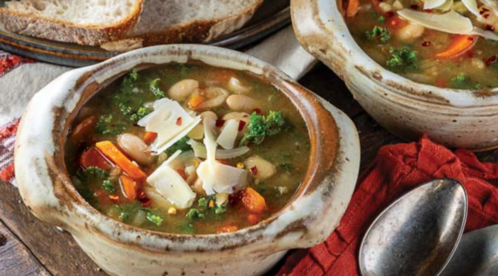 Two rustic bowls of hearty bean and vegetable soup garnished with shaved cheese and red pepper flakes. A spoon rests nearby on a red cloth, and crusty bread slices can be seen in the background.