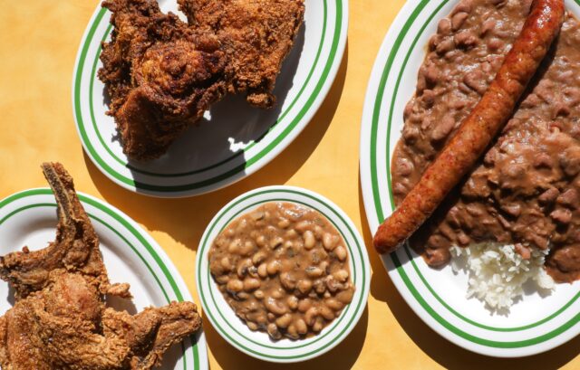 Four plates of food on a yellow table: fried chicken, beans in sauce, sausage with beans and rice, and a bowl of baked beans.