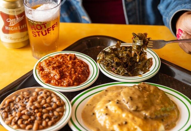 A tray of Southern food including beans, greens, cornbread, and barbecue sauce with a beer glass marked "HOT STUFF" and a Miller High Life can.